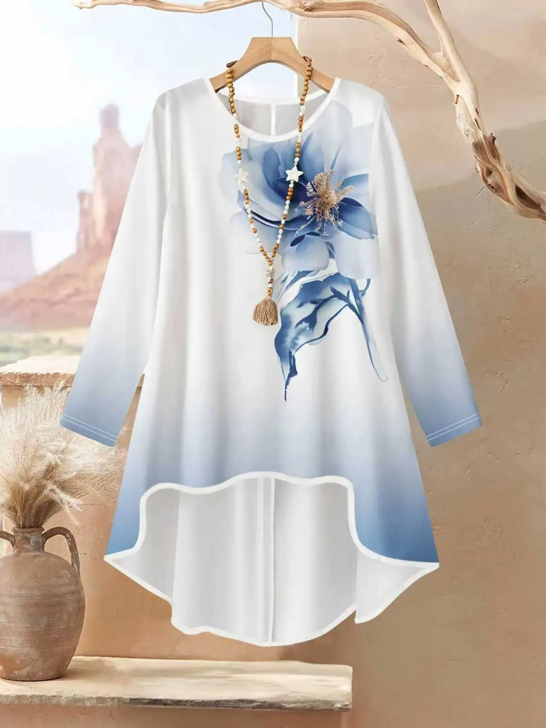 Ein fließendes weißes und blaues Farbverlauf-Langarmoberteil mit großem Blumendruck auf der Vorderseite, das von einem Holzkleiderbügel als Teil eines zweiteiligen Outfits mit Blumendruck und weiter Beinweite hängt.