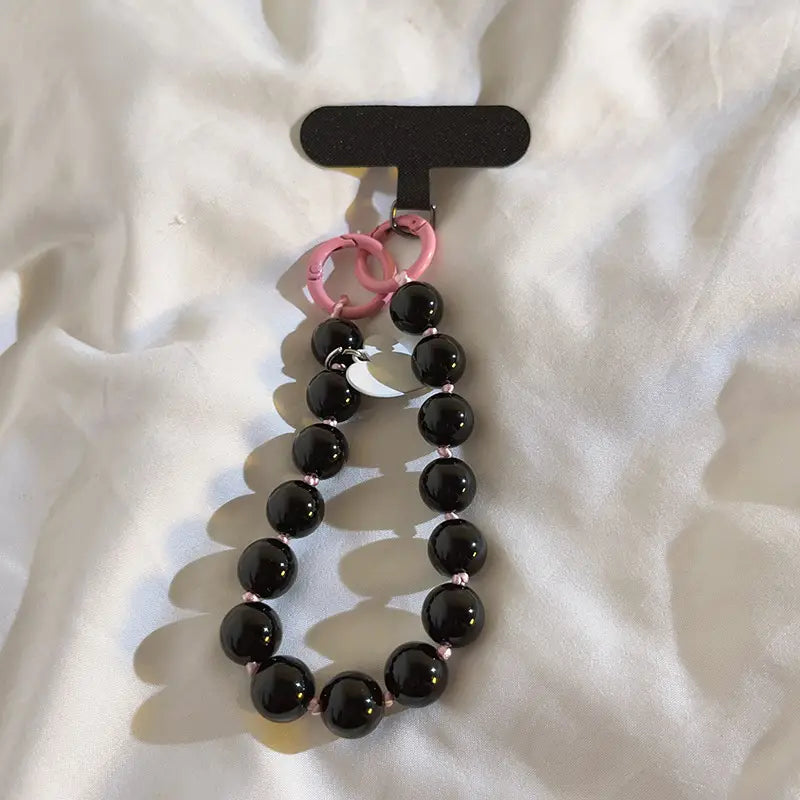 Correa de muñeca para teléfono con cuentas engarzadas que presenta cuentas negras con anillas de plástico rosadas y un dije de luna creciente plateado.