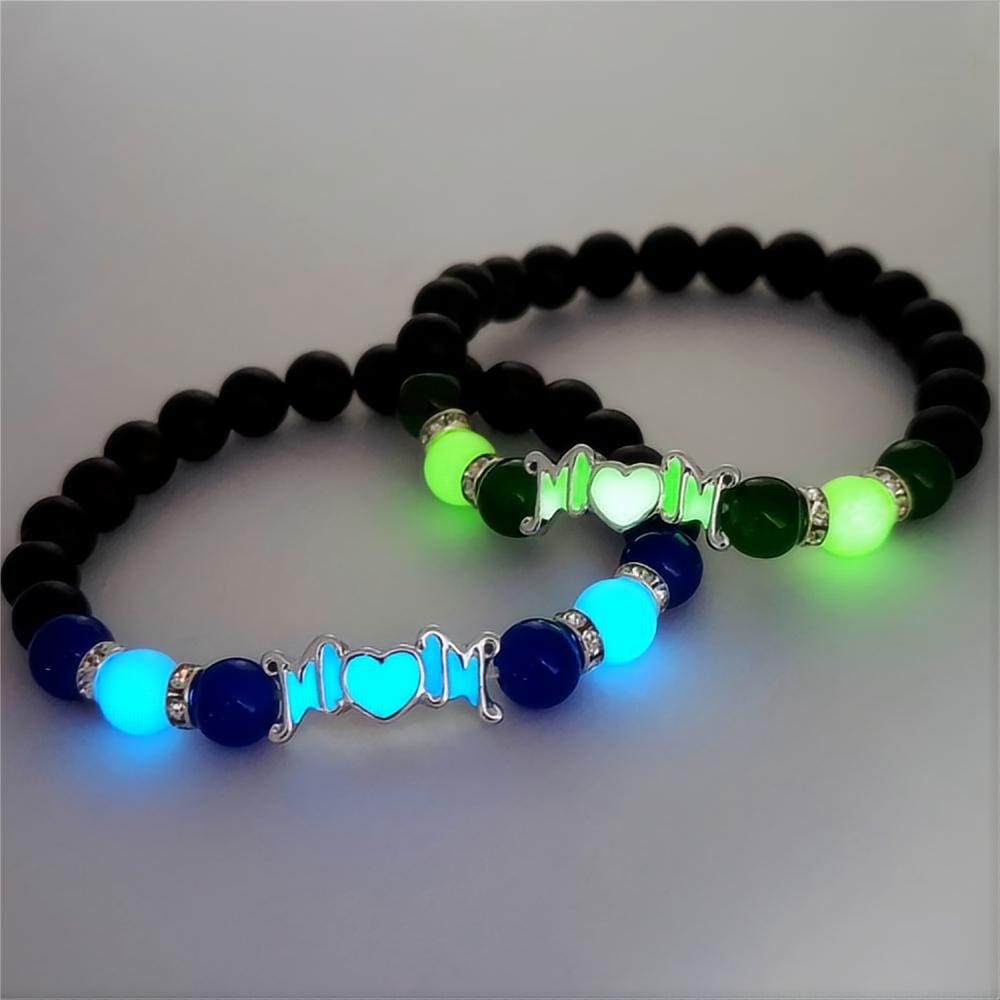 Dos pulseras brillantes con cuentas para madre, una con cuentas negras con acentos verde neón y un dije plateado para mamá y la otra con cuentas azul marino con brillo cian intenso; ambas adornadas con pedrería brillante y diseñadas como regalos de joyería que brillan en la oscuridad.