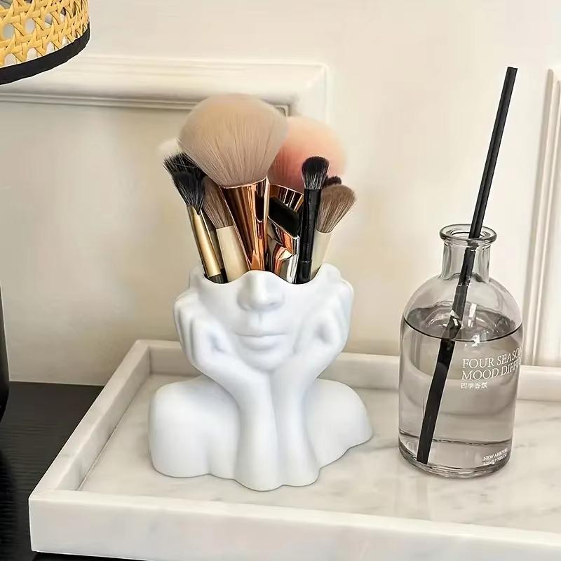 white ceramic abstract face makeup brush holder shaped like a woman’s face with hands resting on cheeks and also functions as a vase