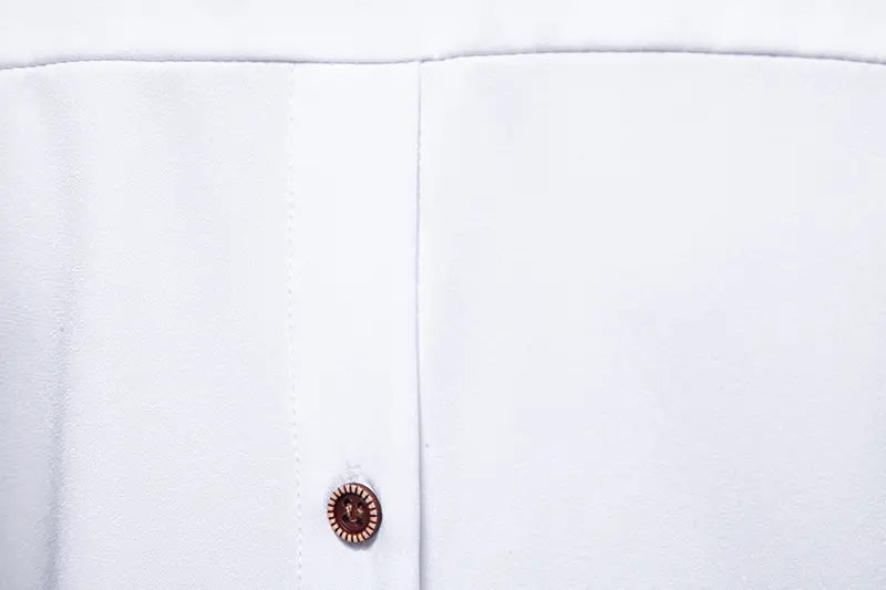 A single dark brown ornate button with a textured circular design on the front of a crisp white men asymmetrical slant collar long sleeve casual shirt.