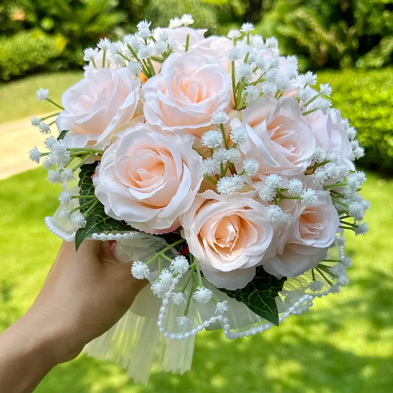 a delicate bouquet of peach-colored artificial roses accented with white baby’s breath and green leaves tied with a sheer lace ribbon in the white silk rose wedding bouquet with lace and pearls collection