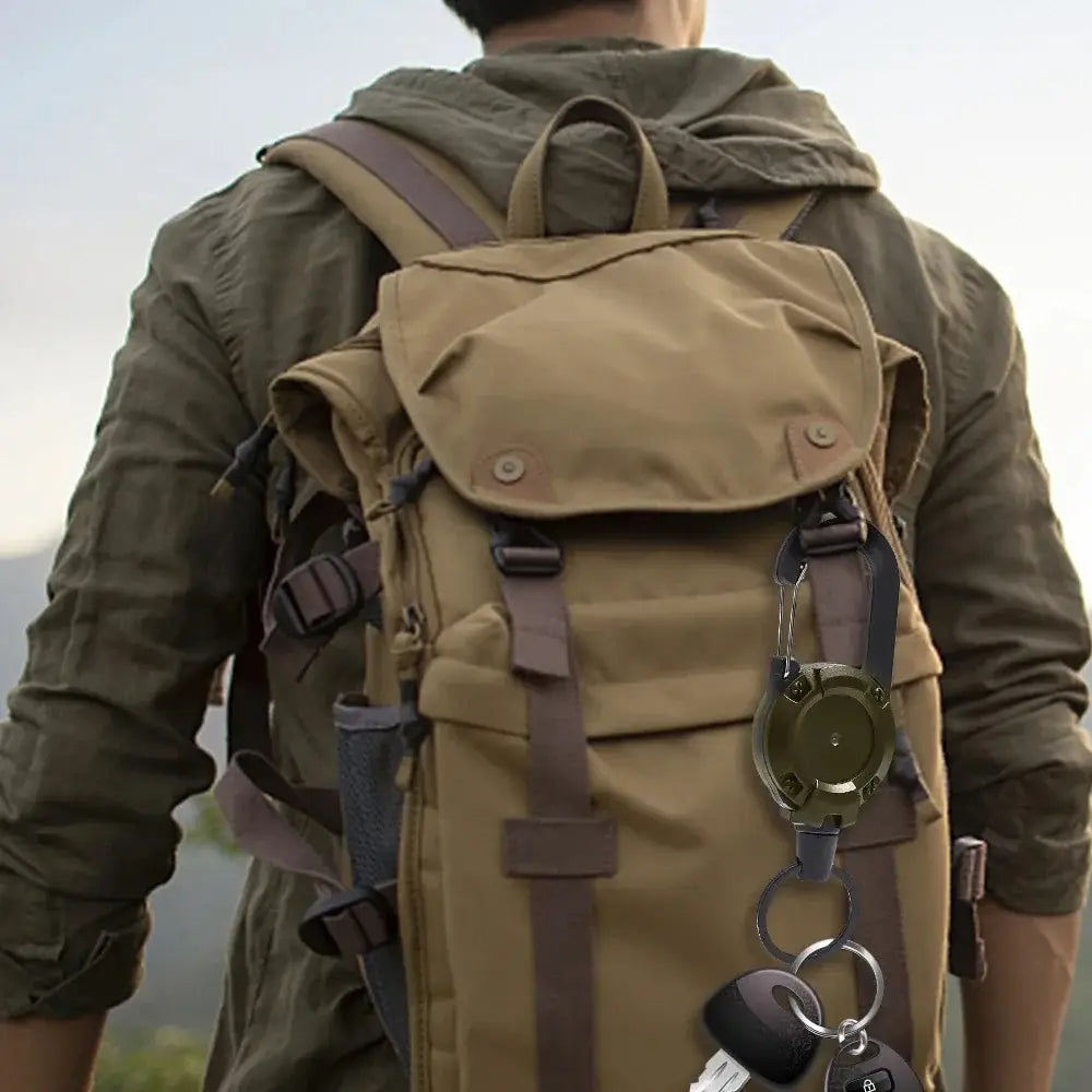 Olive green tactical compass attached to a retractable carabiner keychain made of heavy duty steel wire hanging from a backpack strap.