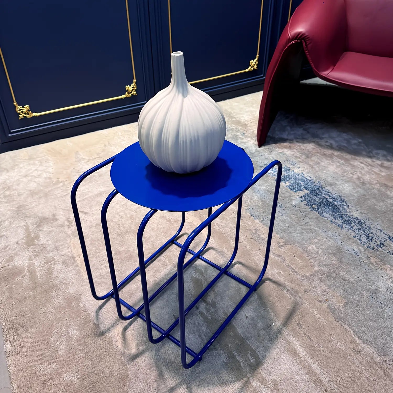 white ceramic garlic-shaped vase displayed on a vibrant blue metal side table with curved frame design featuring minimalist storage and magazine rack functionality