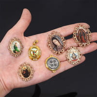 A hand holds a collection of ornate oval-shaped brooches with intricate gold-toned metalwork and varied gemstone inlays alongside a Saint Christopher leather keychain in antique bronze.