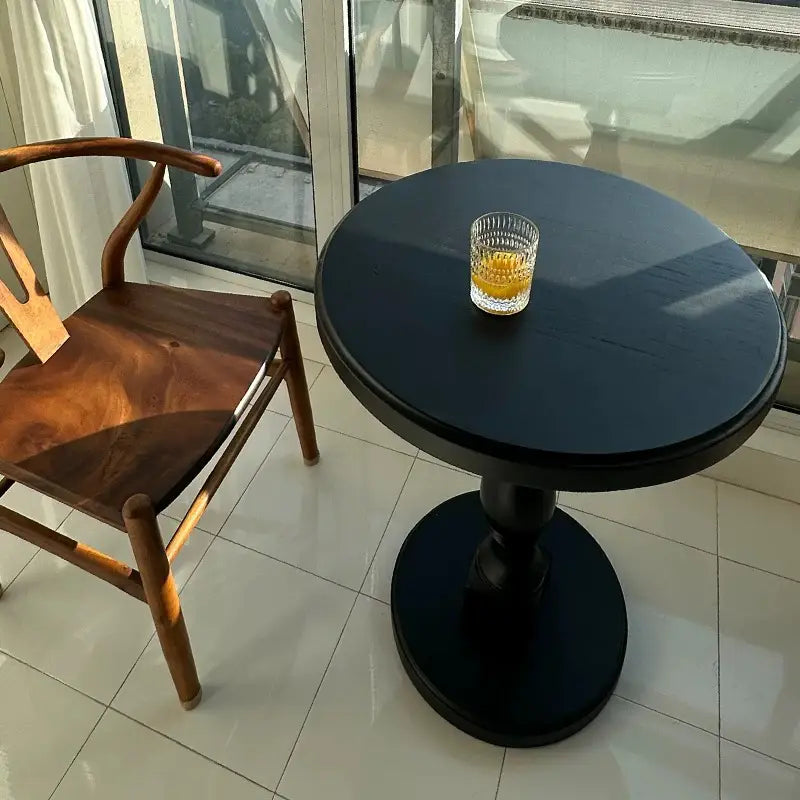 black wooden pedestal round accent table with a glass of amber liquid on top showcasing its black round design and wooden material.