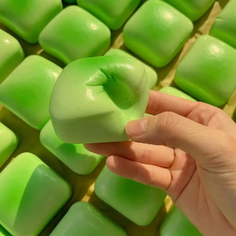 A hand holds a glossy light green mini toast slow rising squishy toy with a square shape and slightly indented center.