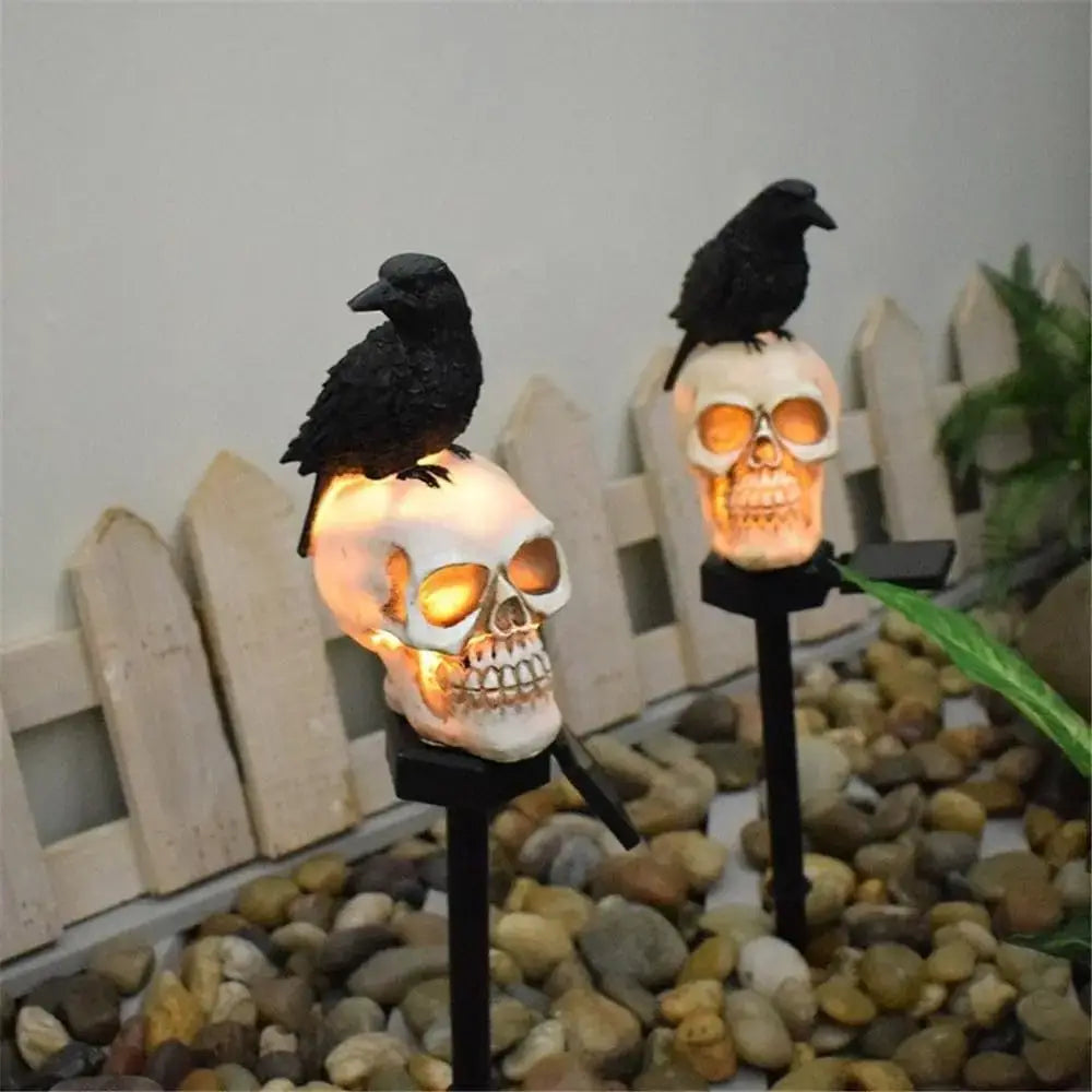 lampe solaire de jardin en forme de crâne et corbeau pour Halloween, avec des lumières de jardin en forme de crâne blanc et des corbeaux noirs perchés au sommet, des yeux orange lumineux, alimentée par un panneau solaire à la base.