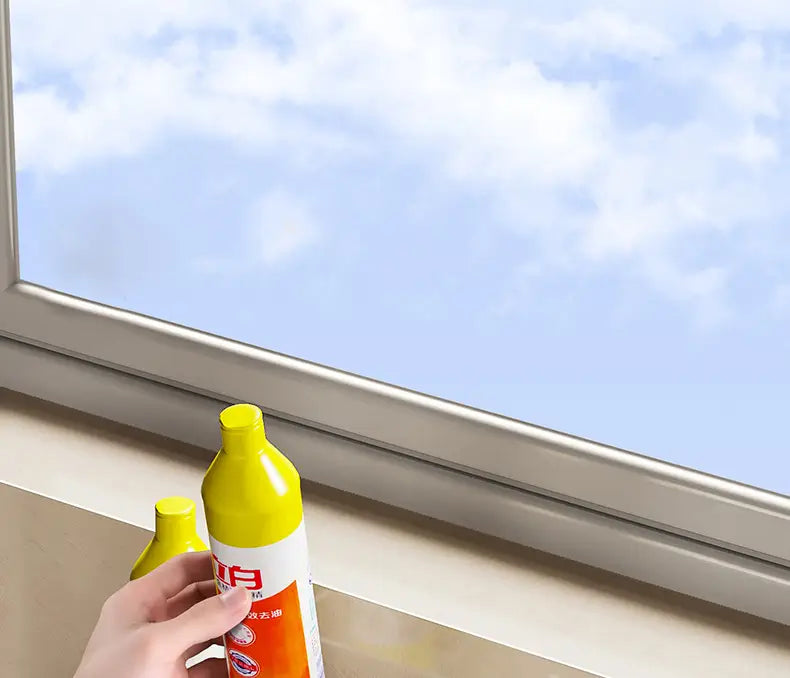 Una botella rociadora con tapa amarilla y etiqueta naranja y blanca, sostenida por una mano cerca de una ventana junto a un organizador para fregadero de cocina de acero inoxidable.