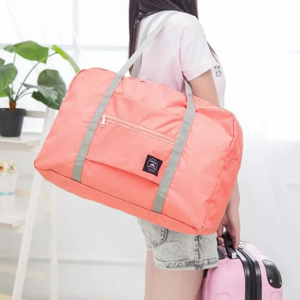 Bolsa de lona de viaje plegable color coral con correas grises y un pequeño parche negro de la marca, diseñada para viajes ligeros.
