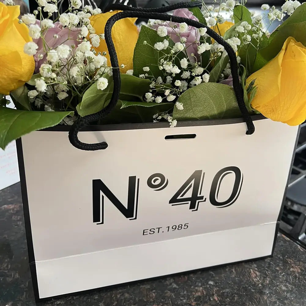 Eine Tragetasche aus weißem Papier mit schwarzen Henkelgriffen und markanter n°40-Kennzeichnung, gefüllt mit gelben Rosen, rosa Blüten und Schleierkraut, gestaltet als Geschenkbeutel zum runden Geburtstag mit Karte und Schleife.