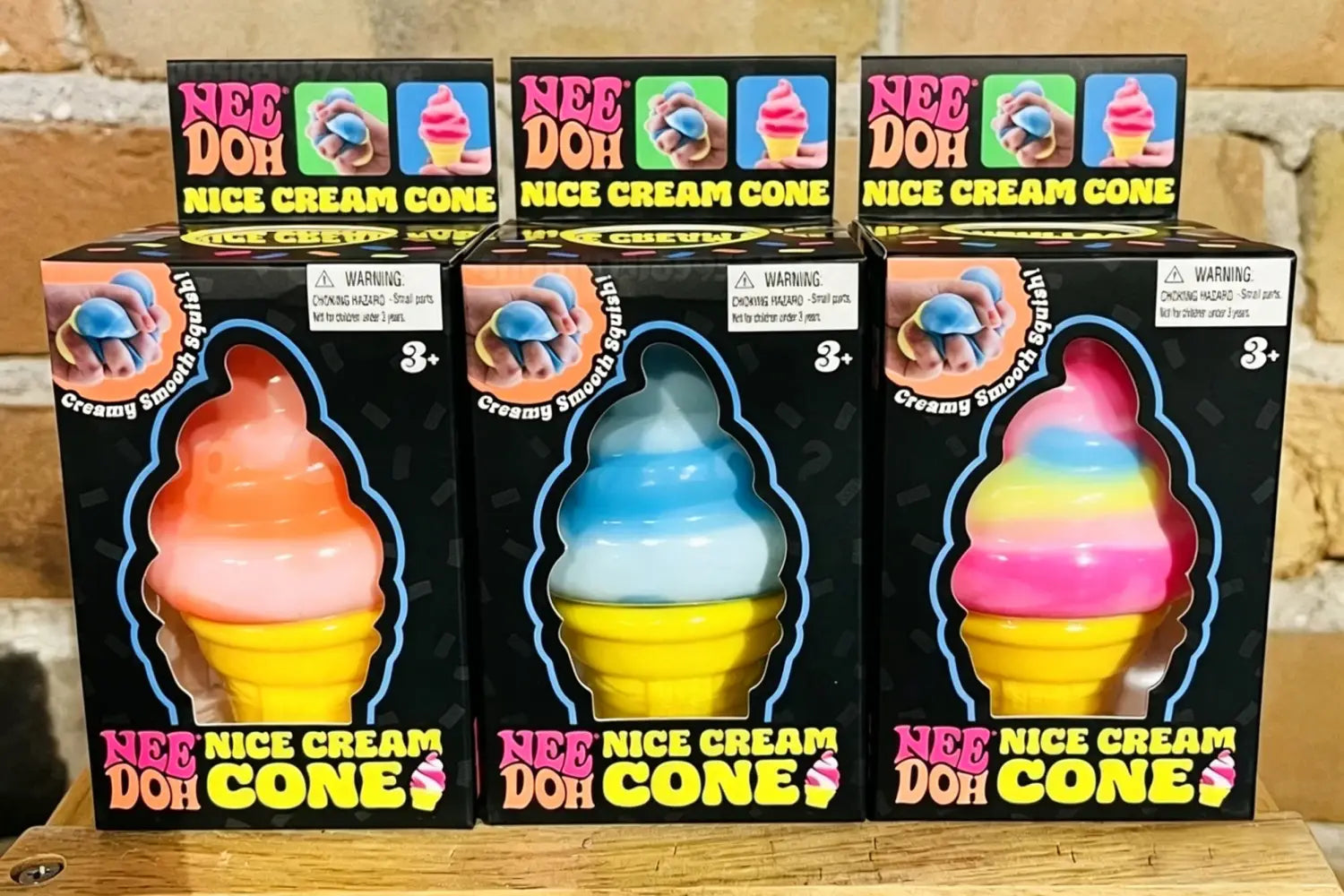 Drei Nee-Doh-Eistüten-Eiscreme-Quietschtier-Fühlspielzeuge mit farbenfrohen geschichteten Eiscreme-Designs in orange-pink, blau-weiß und regenbogenfarbenem pink auf gelben Waffeltüten, von vorne betrachtet.
