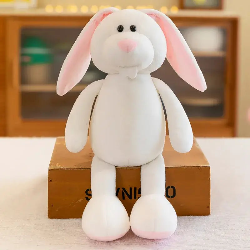 Un animal de peluche conejo relleno blanco de peluche con orejas internas rosadas y una textura suave y acogedora, colocado en posición erguida sobre una caja de cartón en una colección que incluye juguetes blandos de león, elefante y tigre.
