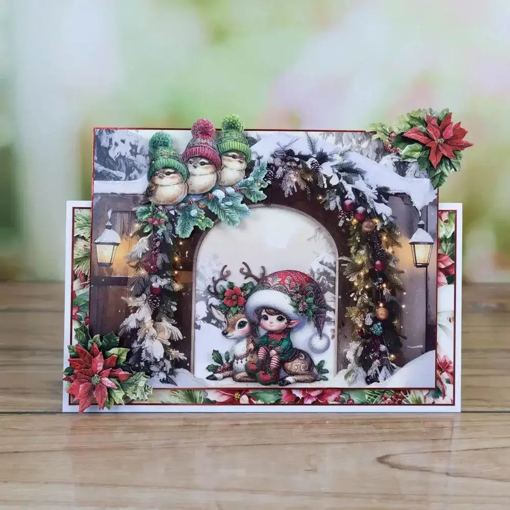 A festive Christmas metal cutting die and stamp set featuring a central illustration of a child in a red and green patterned hat sitting with a reindeer surrounded by snow-covered evergreen branches pinecones and poinsettia flowers.
