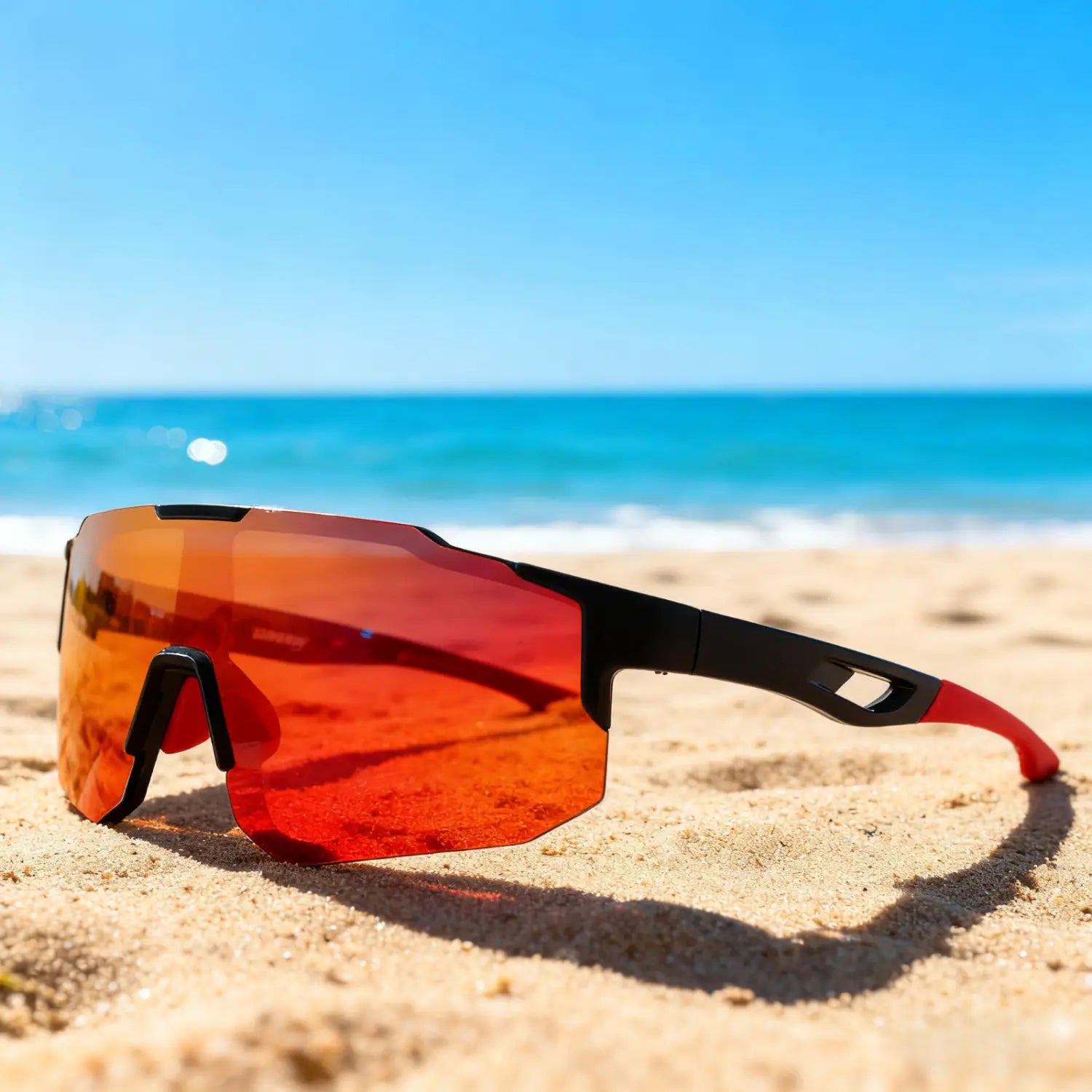 Red and black oversized wraparound sports sunglasses with reflective orange lenses and uv400 protection resting on a sandy beach for unisex comfort fit.