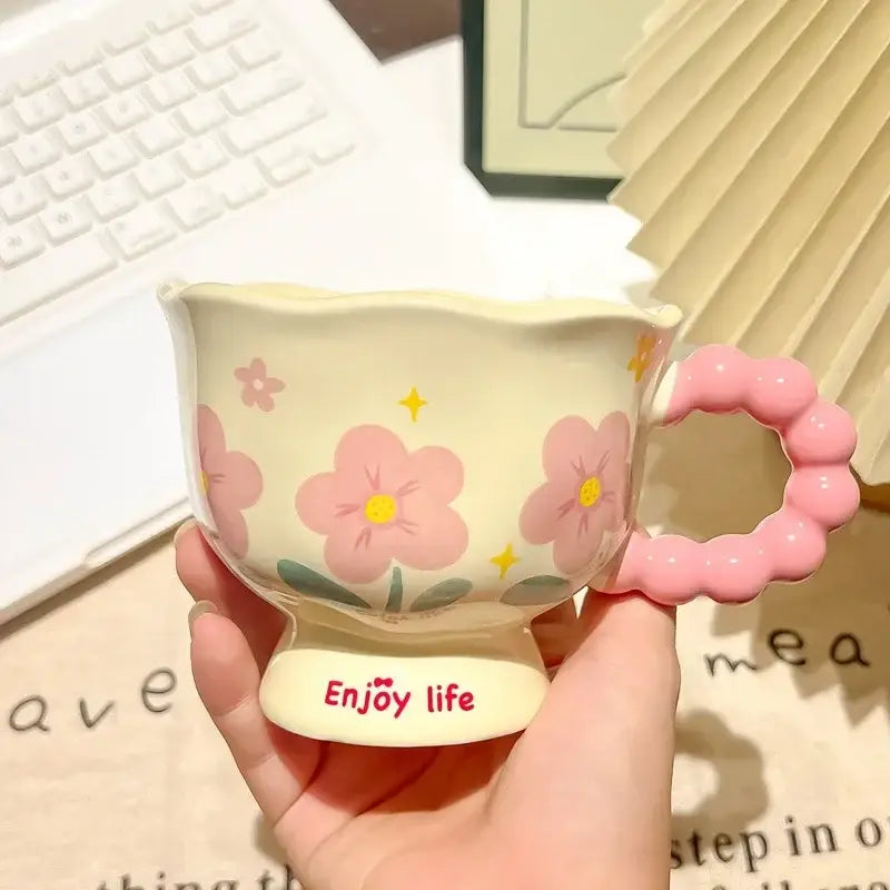 Une tasse tulipe en céramique crème pastel avec des décors floraux roses et une anse rose perlée, portant la mention « enjoy life » en écriture rouge manuscrite sur sa base, dans une boîte cadeau à motif floral.