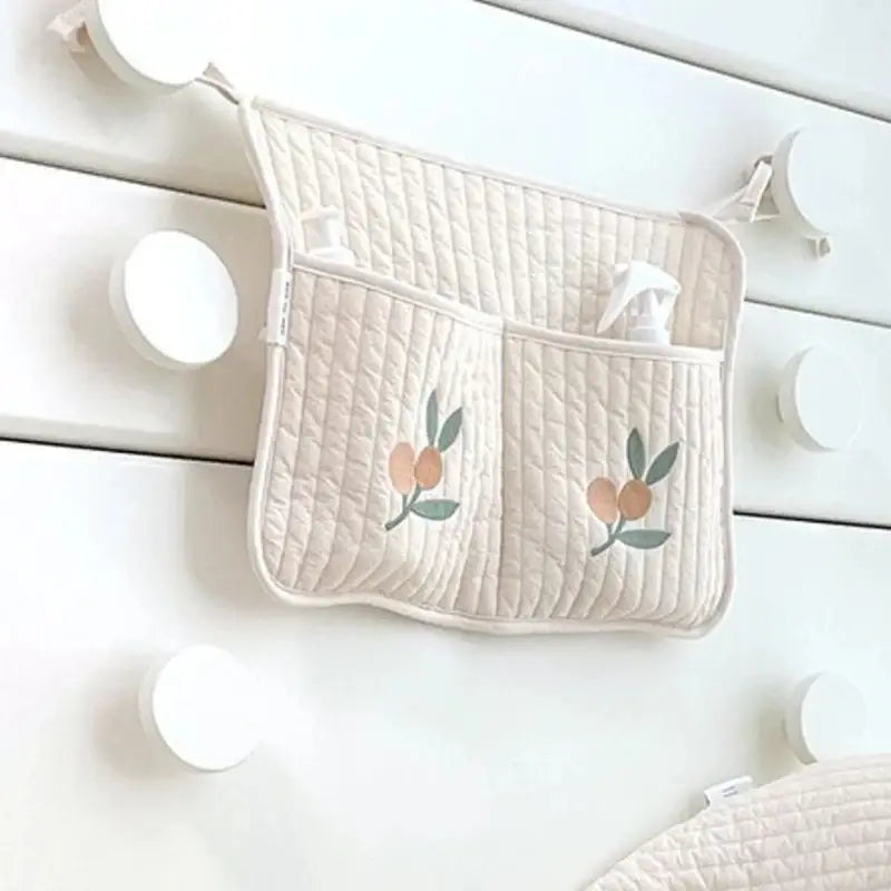 クマの刺繍が施されたクリーム色のキルティング製ベビーカー用オーガナイザー袋で、刺繍の桃と葉のモチーフがあしらわれたポケットが2つ付いています。