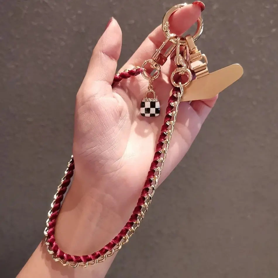 Ein roter geflochtener Kord-Handy-Handgelenkriemen mit goldfarbenen Kettengliedern und einem schwarz-weiß karierten Taschen-Anhänger, entworfen als Anti-Lost-Handylanyard.