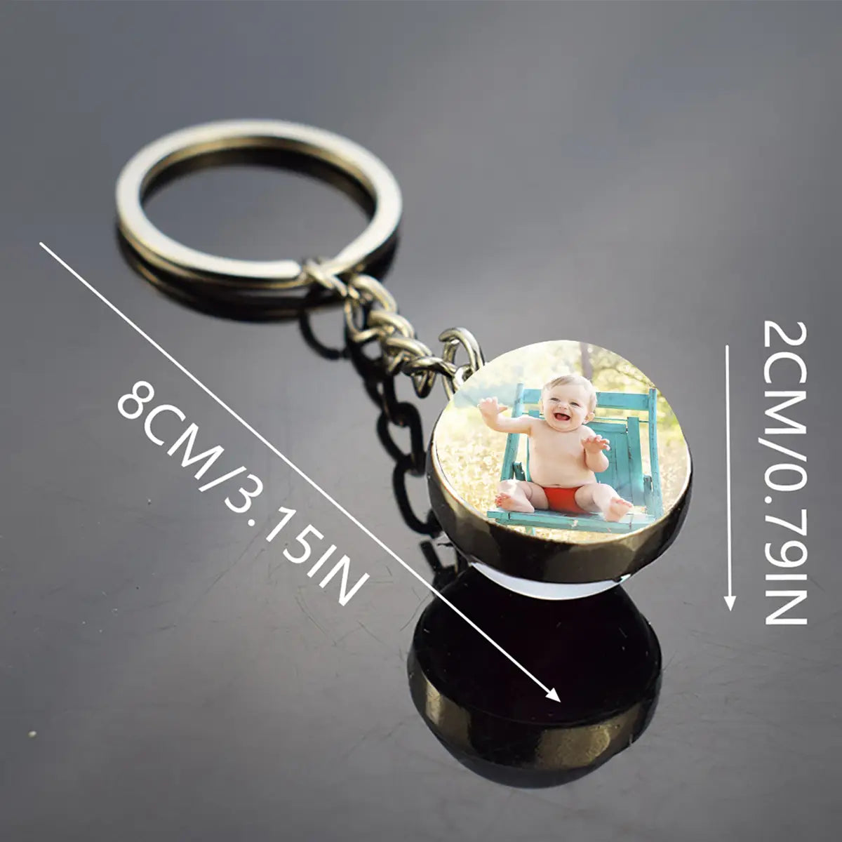 Individuell gestalteter Glühfoto-Schlüsselanhänger, personalisierter runder Schlüsselring, mit einem silberfarbenen Metallring und einer Kette, mit einem glänzenden schwarzen runden Anhänger, der ein gedrucktes Bild eines lächelnden Babys in roten Shorts zeigt, das auf einem blauen Stuhl sitzt.