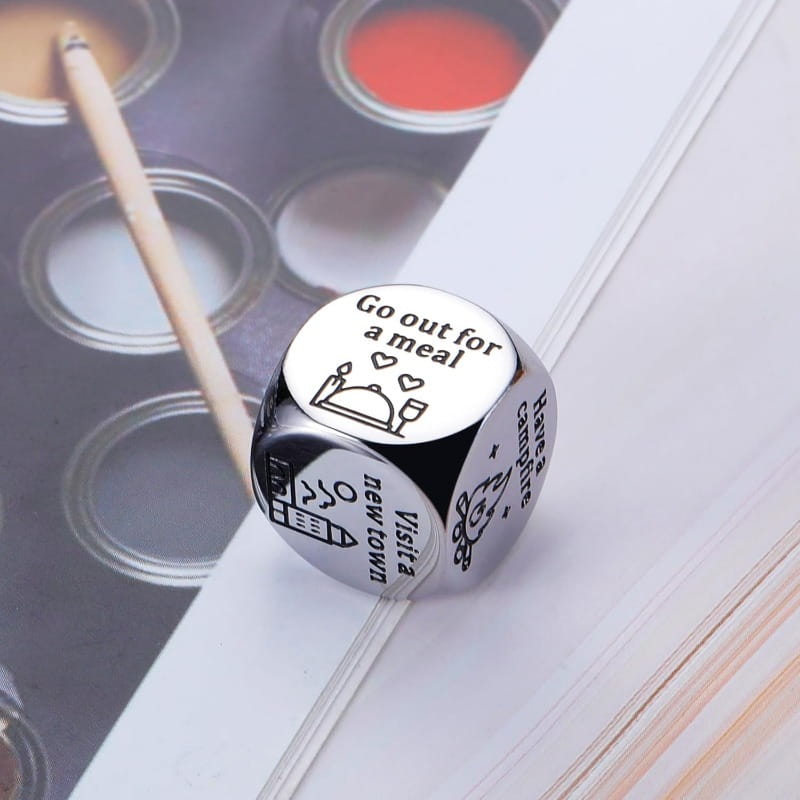 a sleek silver-toned stainless steel six-sided food decision die for couples date night with engraved black text and icons for activities such as go out for a meal and visit a new town