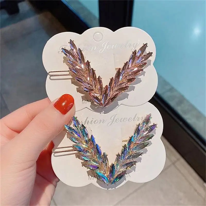 Two iridescent leaf-shaped crystal v shape hair clips in rose gold and blue-green hues adorned with sparkling rhinestones.