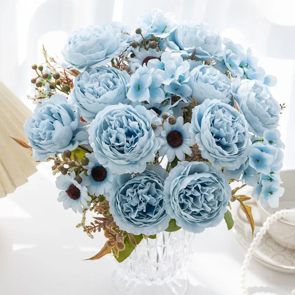 Ein zarter Strauß aus hellblauen künstlichen Pfingstrosen und Hortensien, mit kleinen weißen Gänseblümchen kombiniert, in einer klaren Kristallvase für die Wohnraumdekoration.