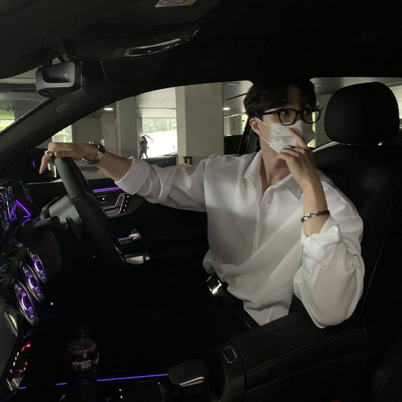 A man wearing a men black long sleeve casual button down shirt with black-framed glasses sits in the driver’s seat of a car with purple ambient lighting.