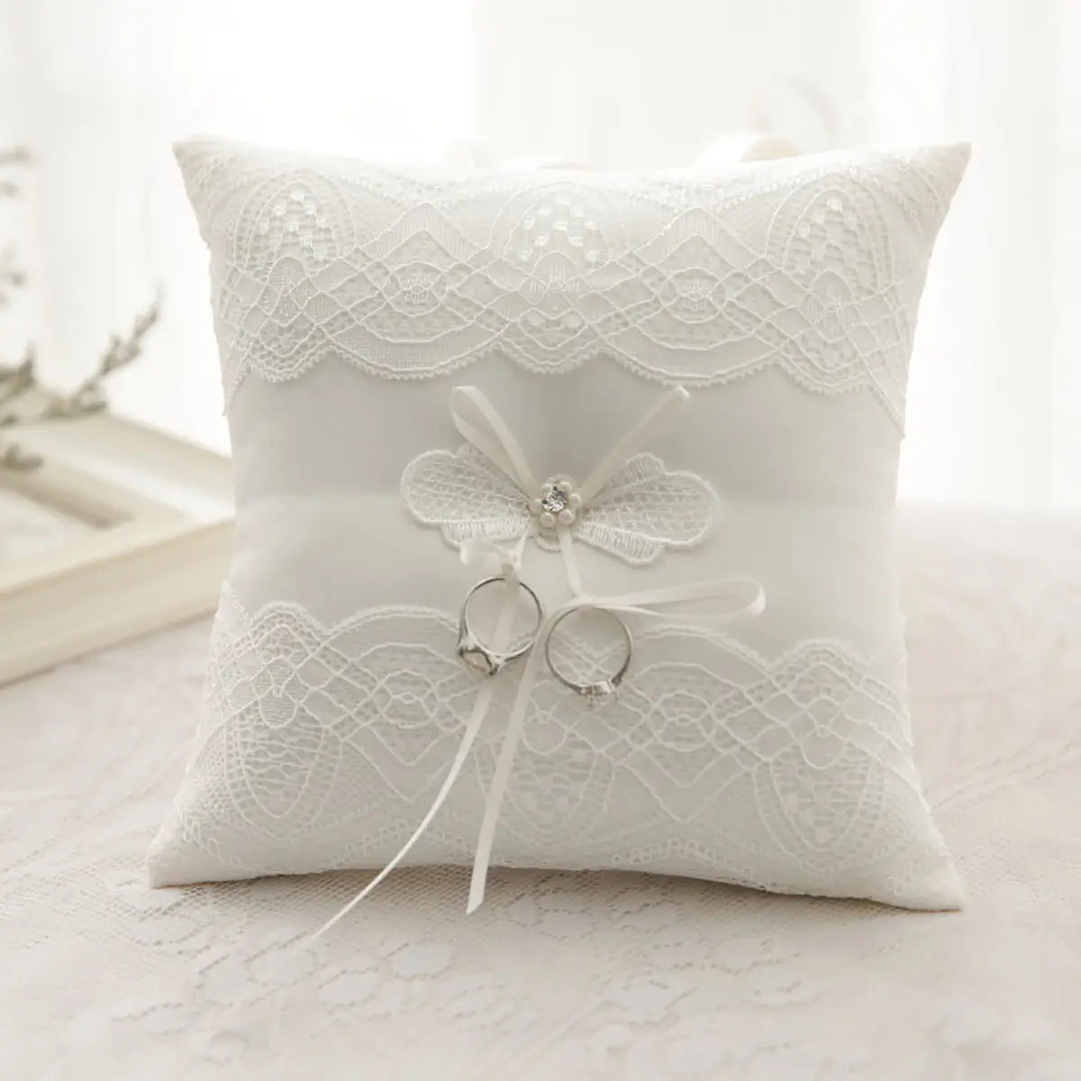Almohada blanca para anillos de boda de encaje con cinta portadora de arras de delicado bordado floral y lazo central adornado con pedrería brillante