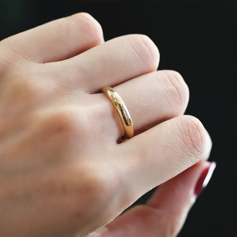 Una elegante argolla de boda de oro de 18 quilates y 4 mm, pulida, con un perfil redondeado suave, llevada en un dedo.