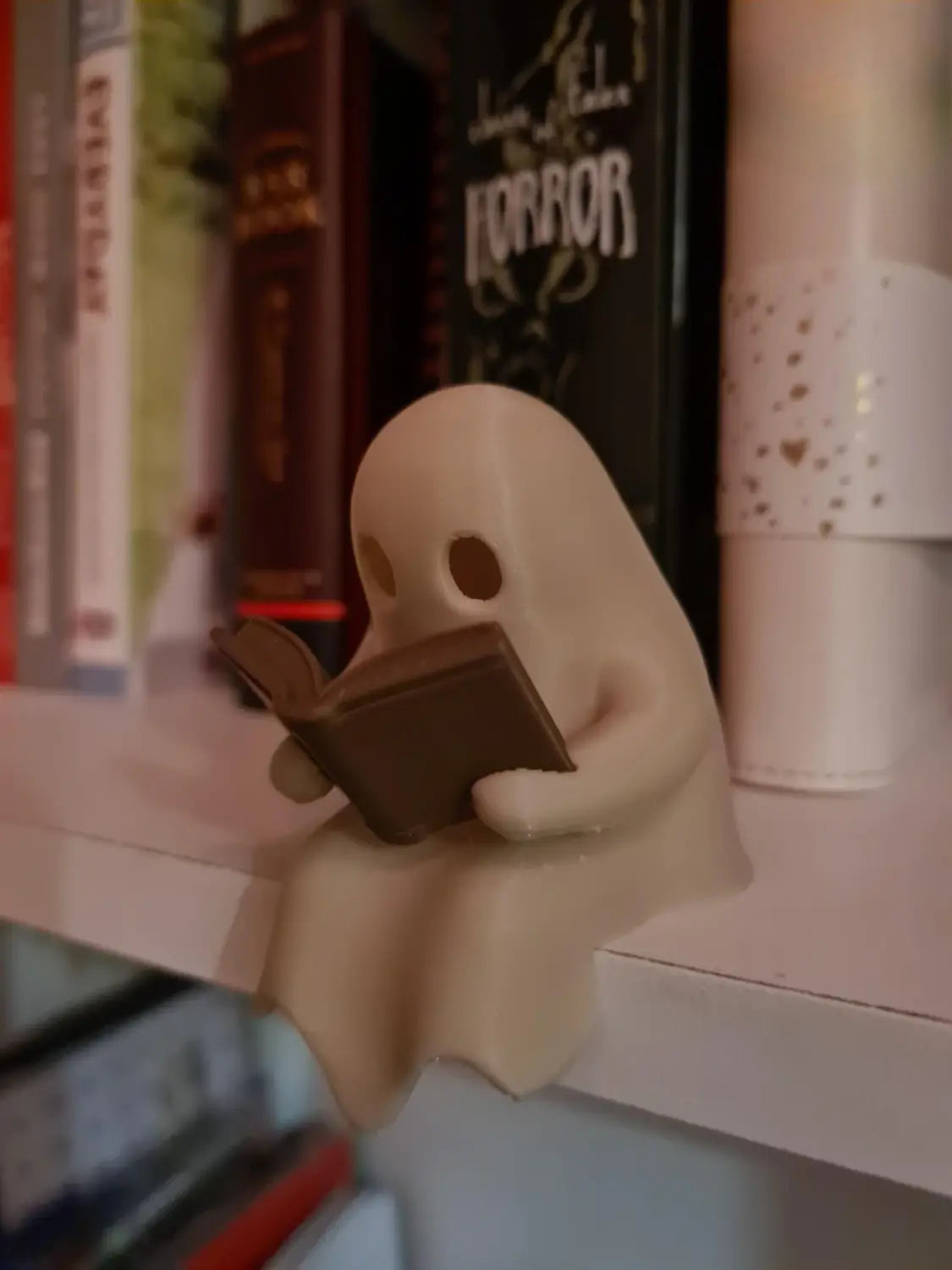 Eine beiges, 3D-gedrucktes Geister-Lese-Buch-Figürchen mit einfachen kreisförmigen Augen und einem fließenden Gewand, das auf einer Ablage sitzt, mit einem flackernden Teelicht.