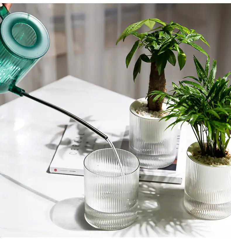 Türkisfarbener, durchscheinender gläserner selbstbewässernder Indoor-Pflanztopf mit schwarzem Auslauf, als Schreibtisch-Planter gestaltet.