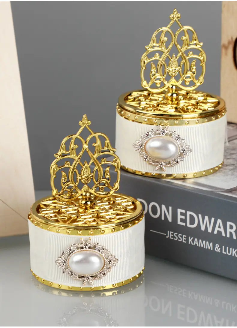 Boîte à bonbons de mariage dorée élégante enveloppée d’un ruban blanc, avec des détails dorés ornés et un ornement de perle centrale, utilisée comme contenant de perle-bonbon décorative.
