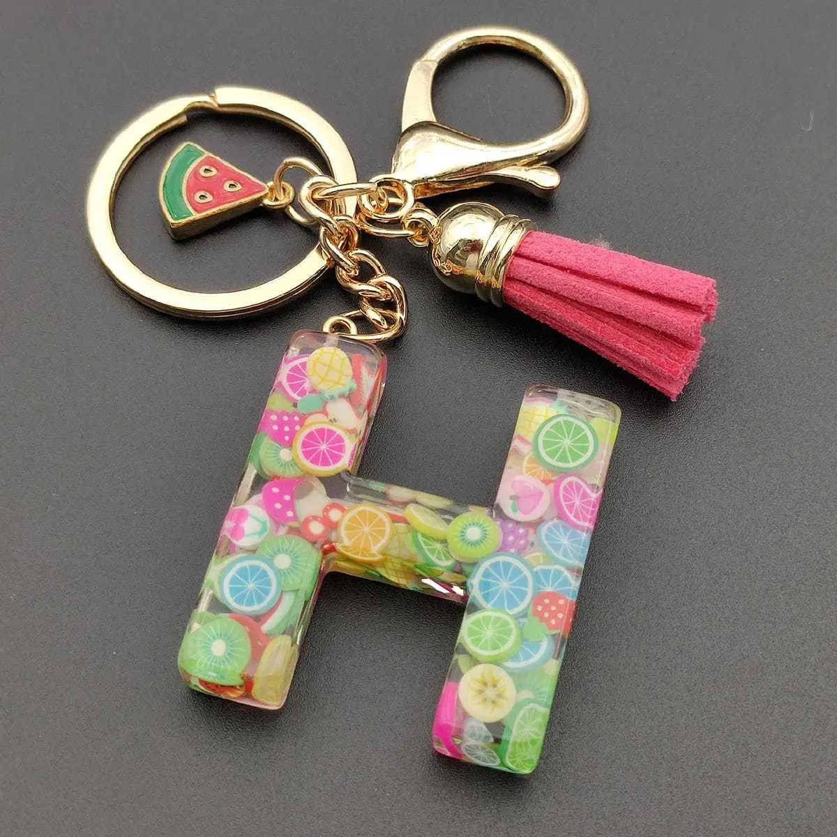 A vibrant transparent resin letter h keychain filled with colorful fruit slice charms accented by a gold toned metal ring clasp and pink suede tassel for a fruit letter resin keychain initial alphabet key ring.