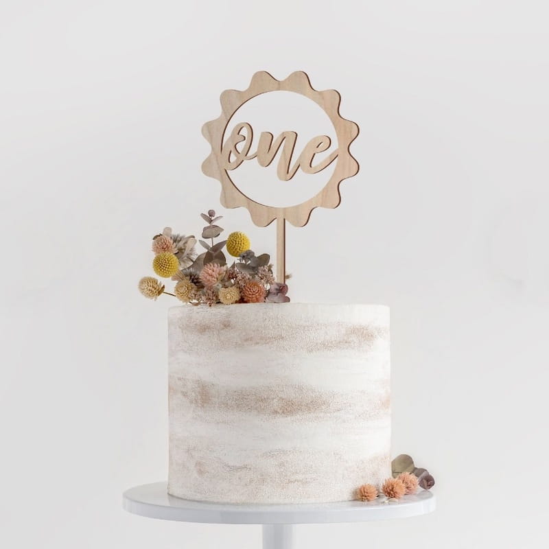 Un délicat topper de gâteau en bois, en forme d’« one », avec un motif en soleil et des fleurs séchées, pour une décoration florale d’anniversaire élégante.