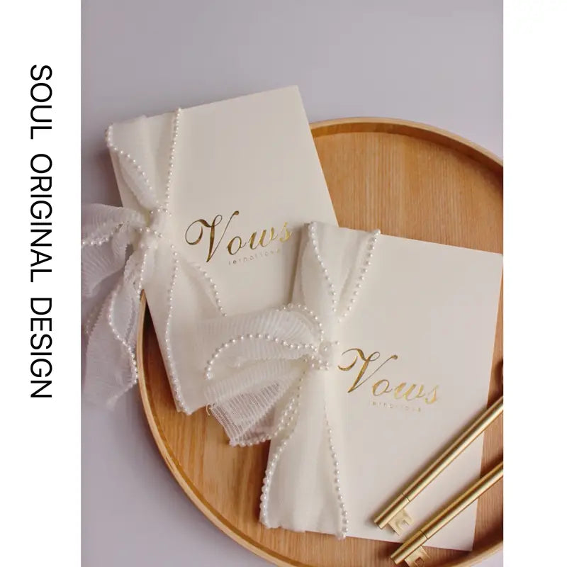 libros de votos de boda color crema con letras con lámina dorada y lazos de cinta semitransparente delicadamente ribeteados con perlas para uso ceremonial elegante.