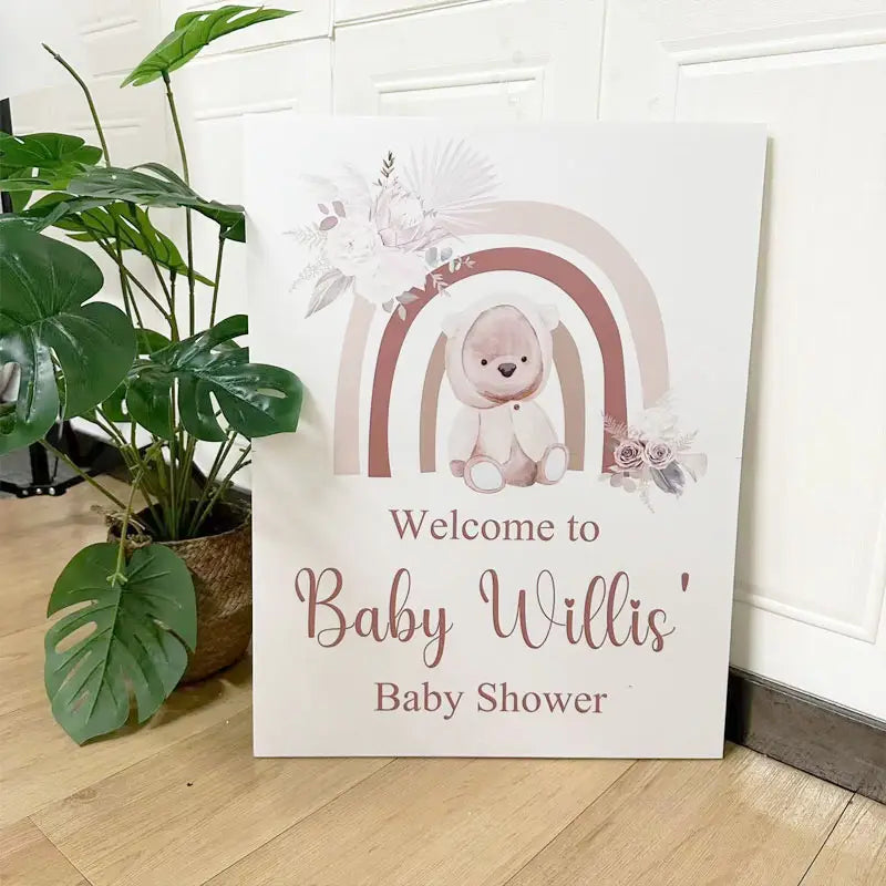 Une enseigne de bienvenue personnalisée pour baby shower blanche et rose pâle, avec un ourson illustré sous un arc-en-ciel, avec des détails floraux conçus pour la décoration de baby shower.