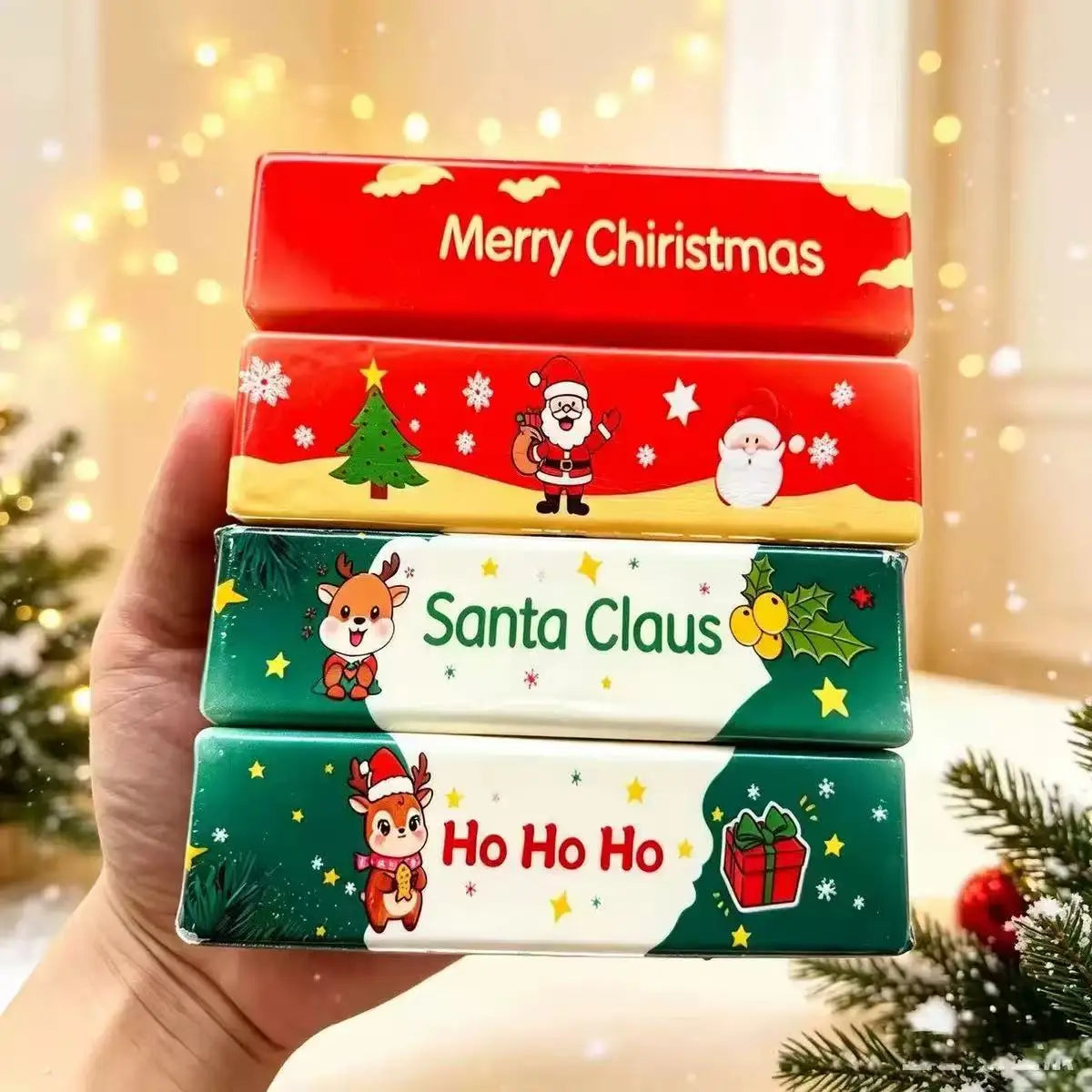 Una mano que sostiene un conjunto de cuatro juguetes de mantequilla blanda y esponjosa de forma rectangular, con un diseño festivo, de descompresión lenta, tipo caja de pañuelos, con diseños de caricaturas coloridas y texto de temporada navideña.