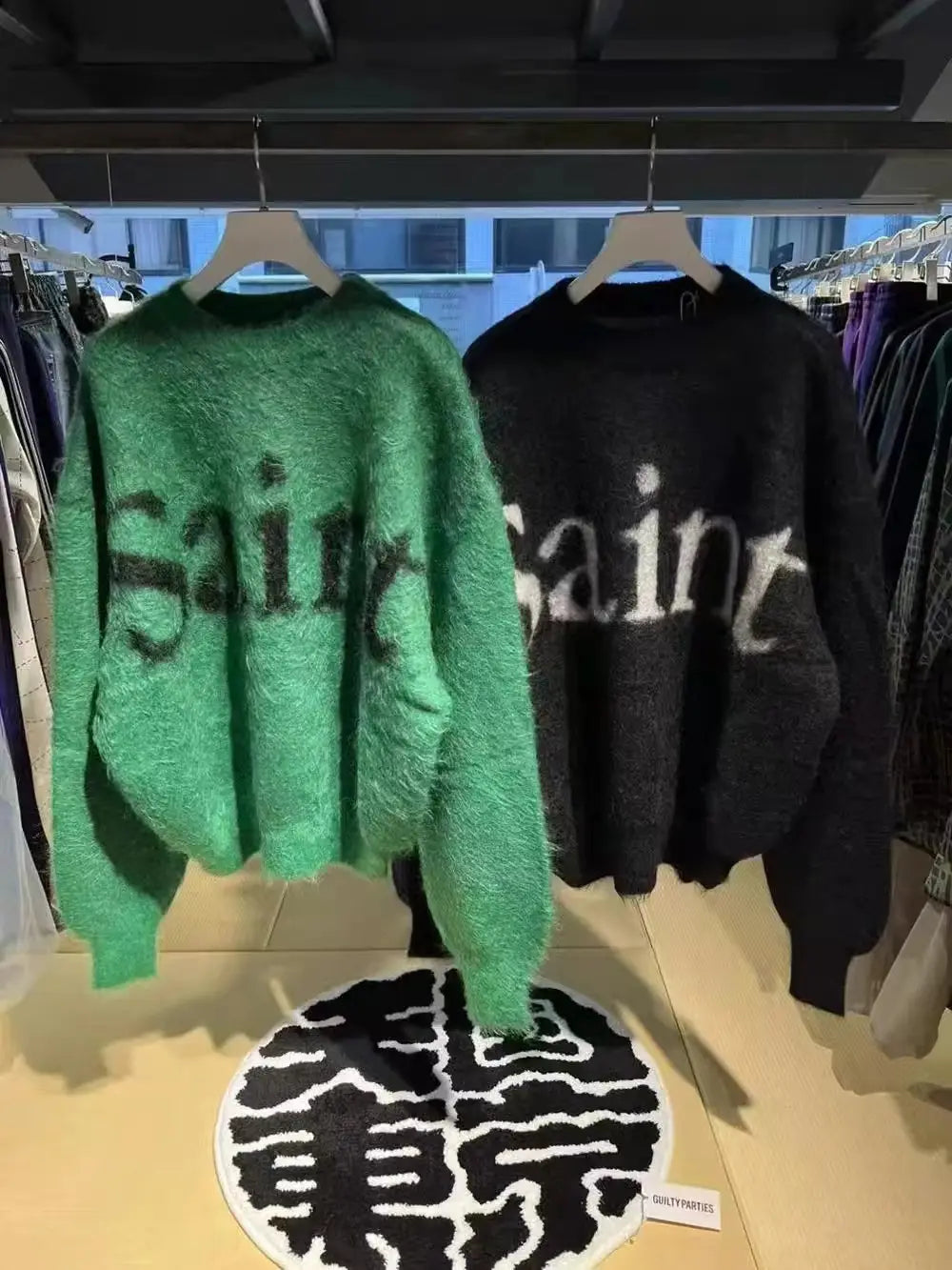 sin刺繍のブラックのオーバーサイズニットセーター、ならびに聖人のレタリングを差し色であしらった緑および黒のフワフワニットセーター（木製ハンガーに掛けられています）
