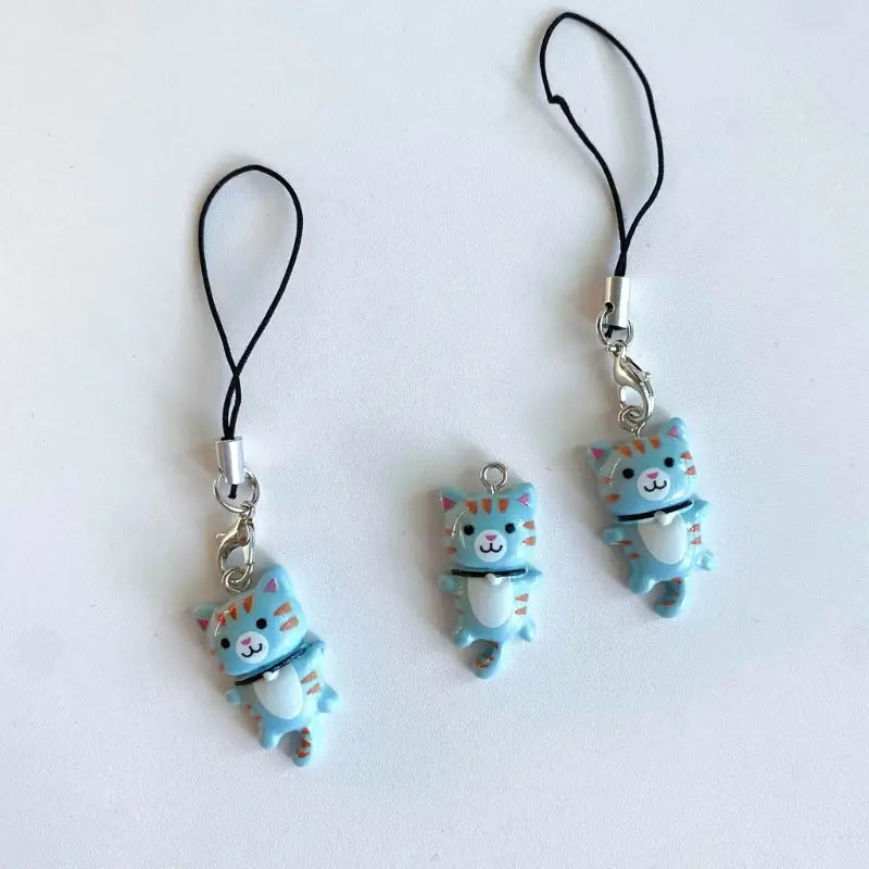 Light blue cat-shaped phone charm strap with orange stripes and white belly attached to black cord with silver metal clasp.