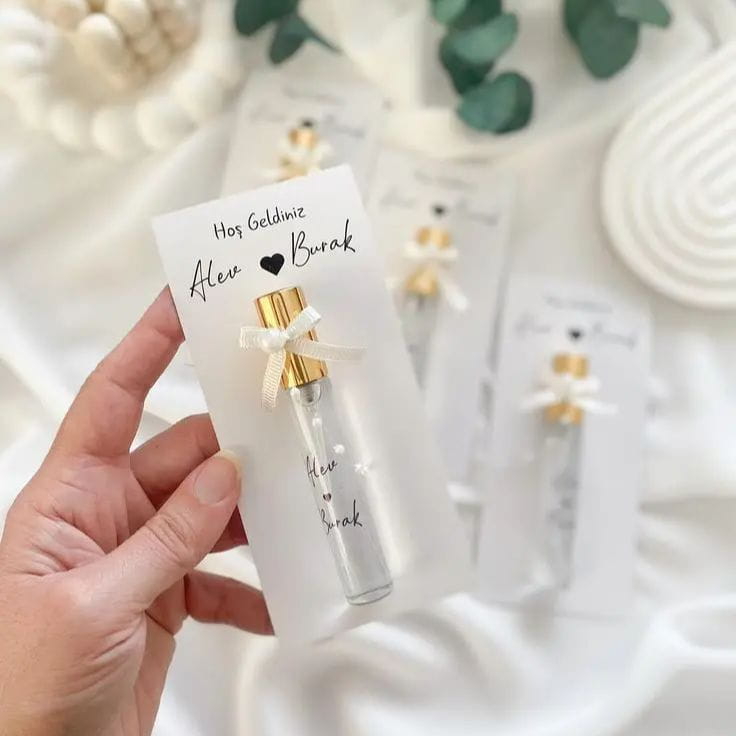 bouteille en verre transparent pour carte cadeau de parfum de mariage personnalisée, avec un capuchon doré et un délicat ruban blanc noué autour de son goulot, offerte dans une carte de faveur personnalisée