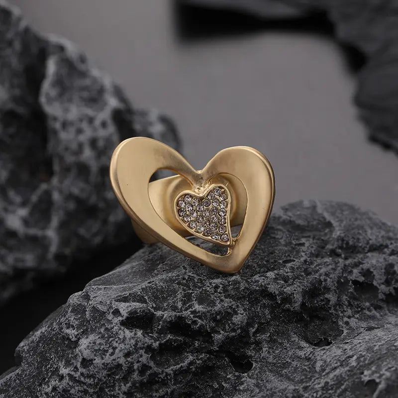 Anillo con forma de corazón ajustable en tono dorado con diseño en capas, que presenta una inserción central de pedrería transparente, ajustable para un uso versátil de joyería.
