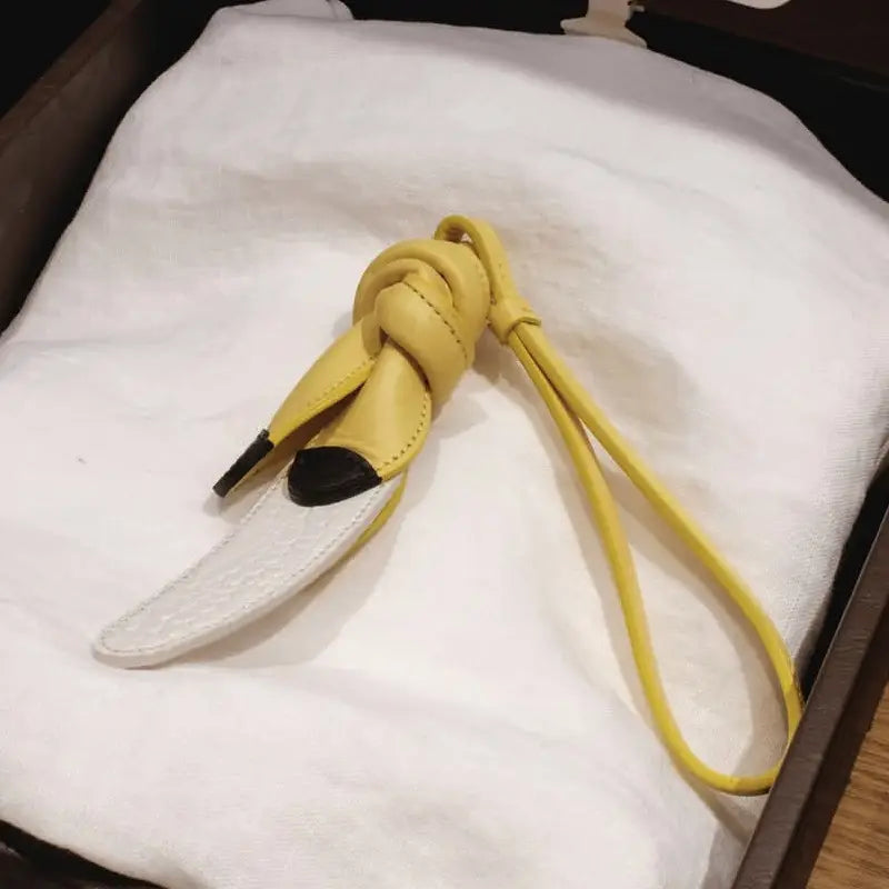 Un porte-clés en forme de banane en cuir jaune, avec des extrémités à pointe noire et une base blanche texturée, conçu comme charm pour sac à main en cuir végétal et ornement de sac à main de créateur.
