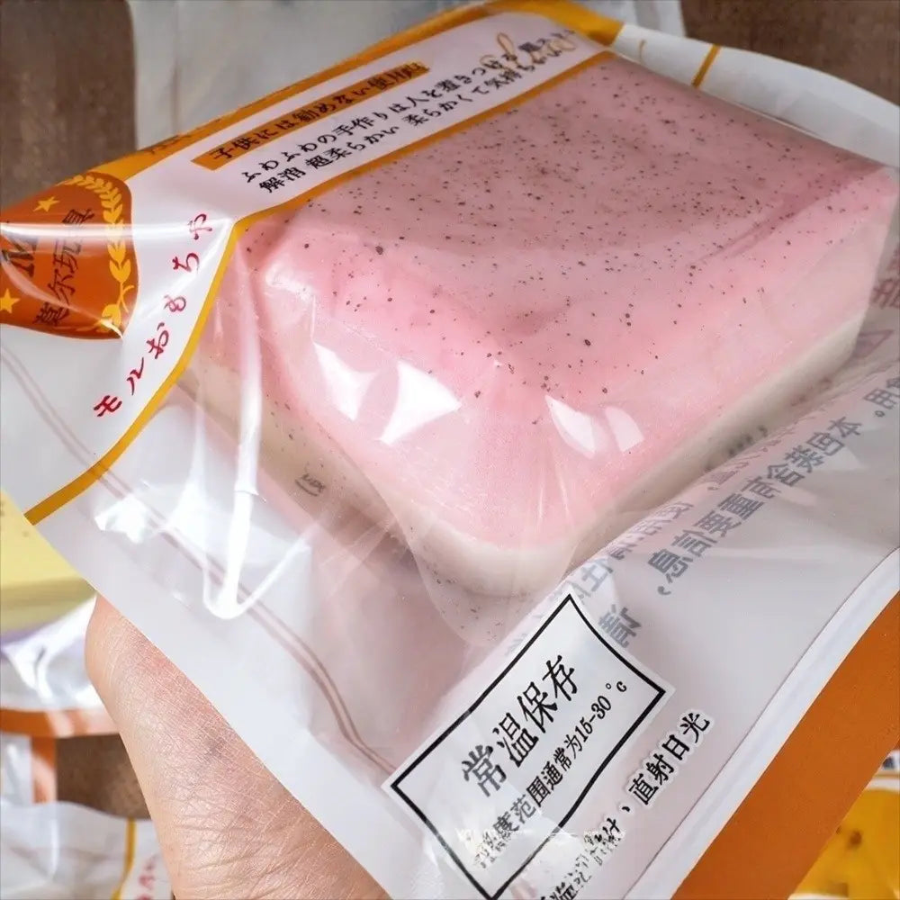 ein pink gesprenkeltes Mochi-Dessert in einer transparenten Plastikhülle mit orangefarbener und weißer Markenkennzeichnung, das neben einem Polyester-Acryl-Winterschal platziert ist