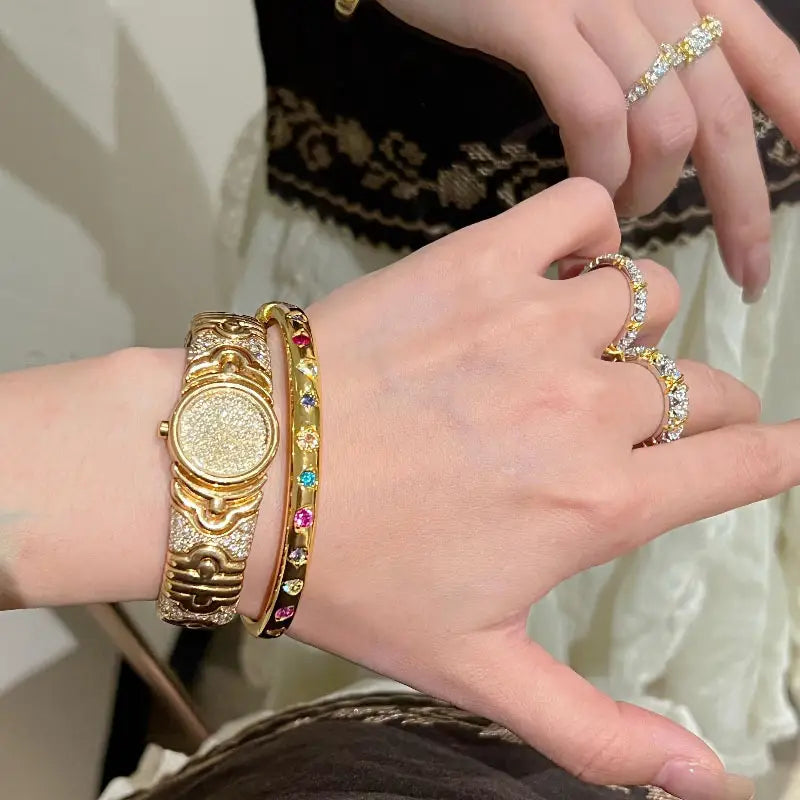 Eine goldfarbene Uhr mit einem strukturiert funkelnden Zifferblatt und aufwendig gestalteter Gehäusekonstruktion, kombiniert mit einem Armreifarmband aus goldfarbenem Multigemsteinschmuck.