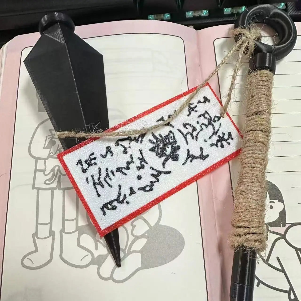 schwarzer, handgefertigter Stift mit strukturierter, mit Jute umwickelter Griffpartie und schwarzer Kunststoffkappe, neben einem Ninja-Kunai-Kühlschrankmagneten aus PVC, Shuriken-Spielzeug.