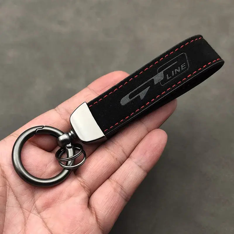 Schwarzer Lederschlüsselanhänger der GT Line aus Wildleder mit roter Steppnaht und einem silbernen Emblem, das einen Metallring und eine Klammer zeigt.