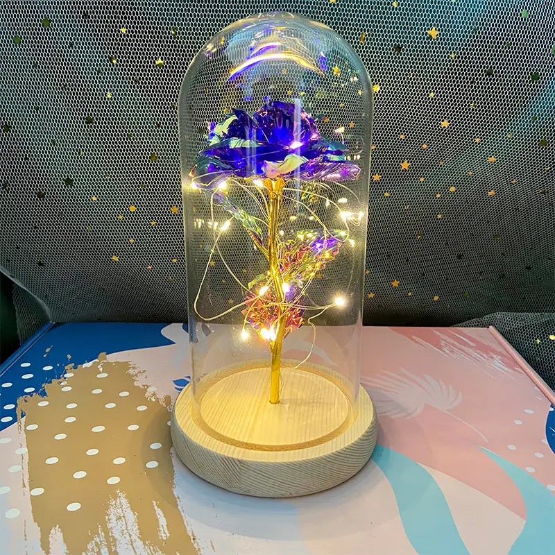 Eine leuchtend violette und goldene LED-beleuchtete Rosentrieb-Optik, umschlossen in einer klaren Glaskuppel, mit Holzsockel, im „Eternal Gold Rose Glass Dome Led Light Gift“.