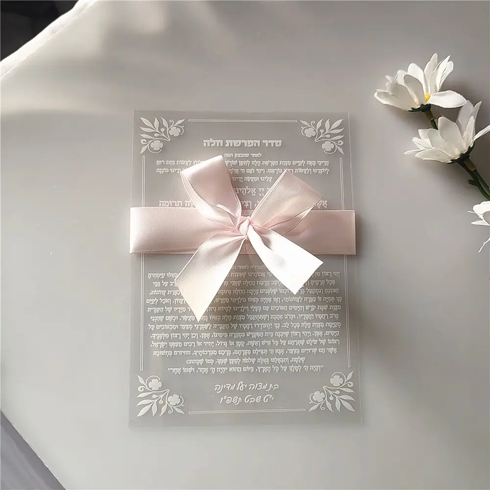 A delicate translucent custom Hebrew acrylic prayer card adorned with a soft pink satin bow and intricate floral etchings designed as a wedding and bar mitzvah souvenir.