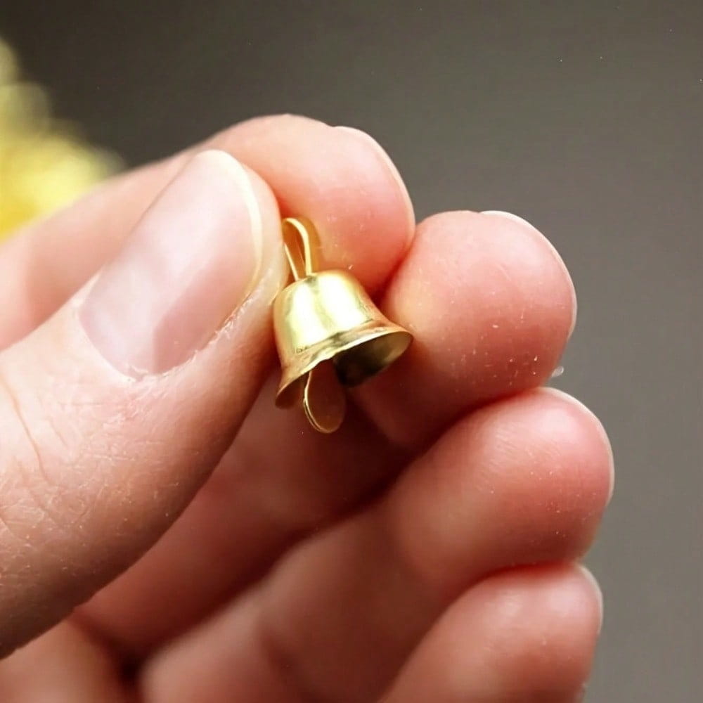 Ein kleiner Anhänger in Mini-Glockenform aus goldenem Material mit polierter metallischer Oberfläche und einer geschlossenen, geschwungenen Schlaufe zur Befestigung, von vorne betrachtet.
