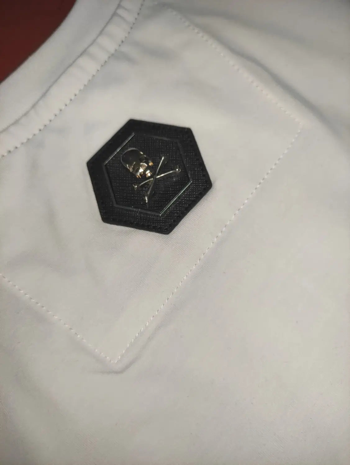 Men rhinestone devil bear graphic tee featuring a black hexagonal patch with a silver skull and crossbones emblem stitched onto white fabric.